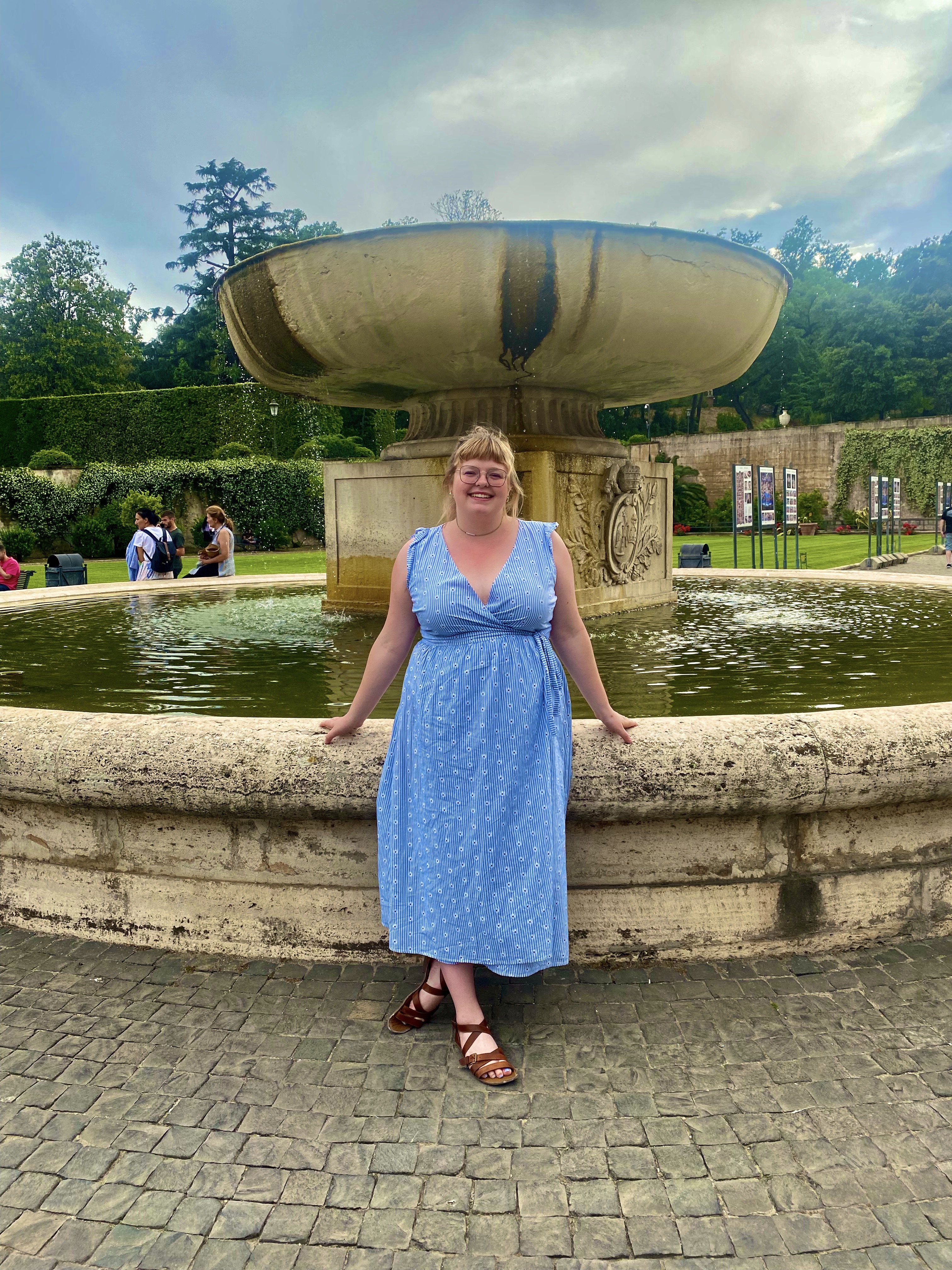 A woman in a blue dress posing in front of a fountain and smiling.